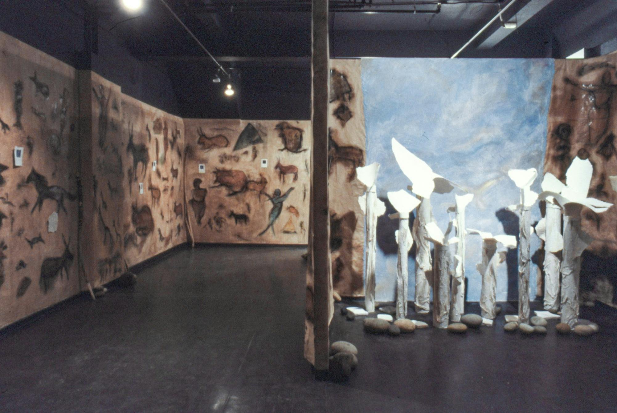 The walls of a room are lined with large chalky drawings of animals and humans. One section is divided and has a large blue patch painted as sky, tall white sculptures, and many rocks on the floor.