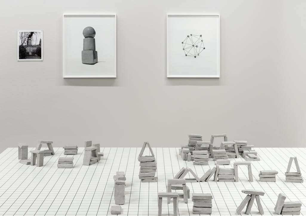 Black and white image of thin clay blocks stacked and grouped on a table. Behind hang framed photographs of simple toy-like structures: stacked blocks and a sphere of connected toothpicks. 