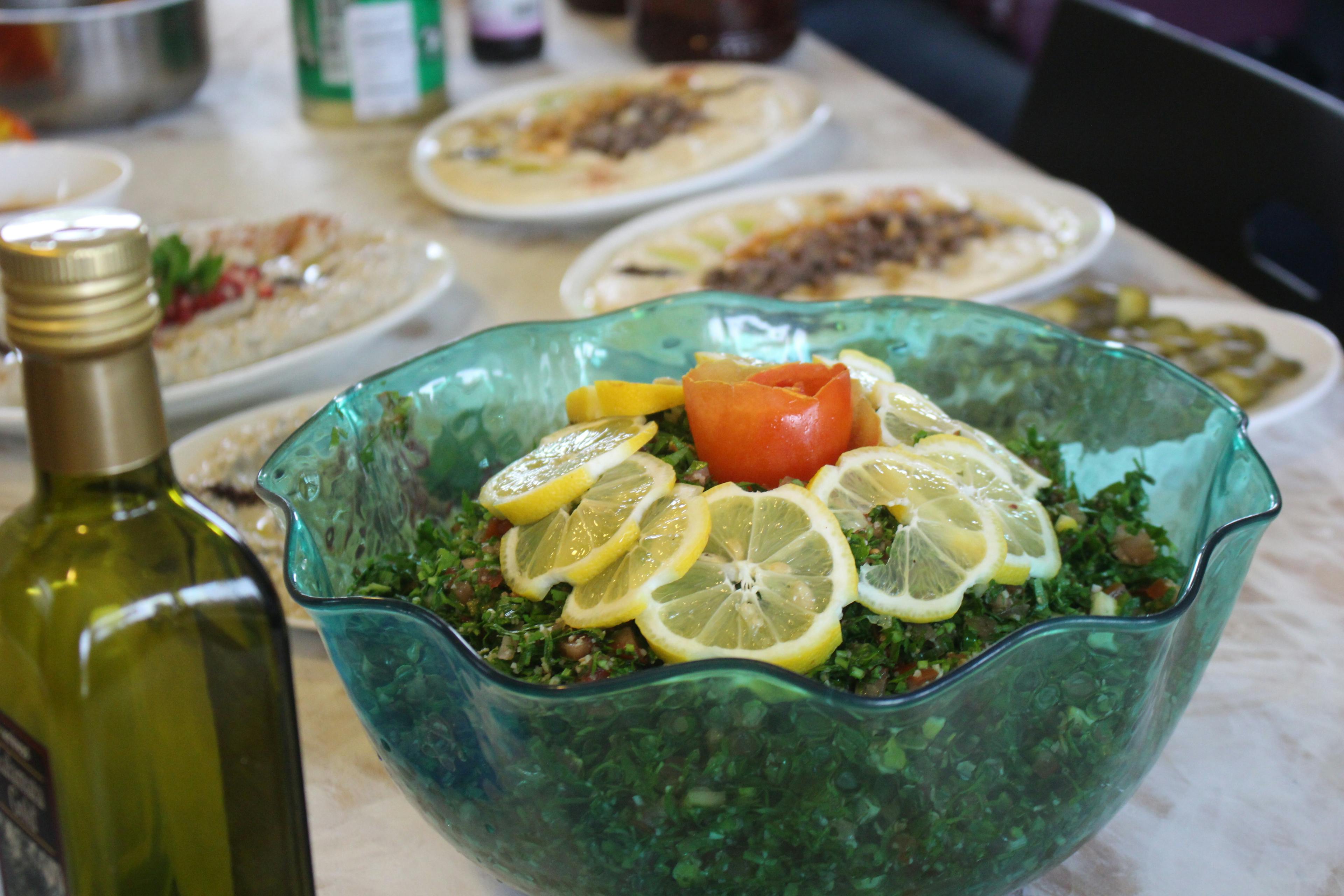 A salad in a big blue bowl, a bottle of olive oil, and various other plates are placed on a table. 