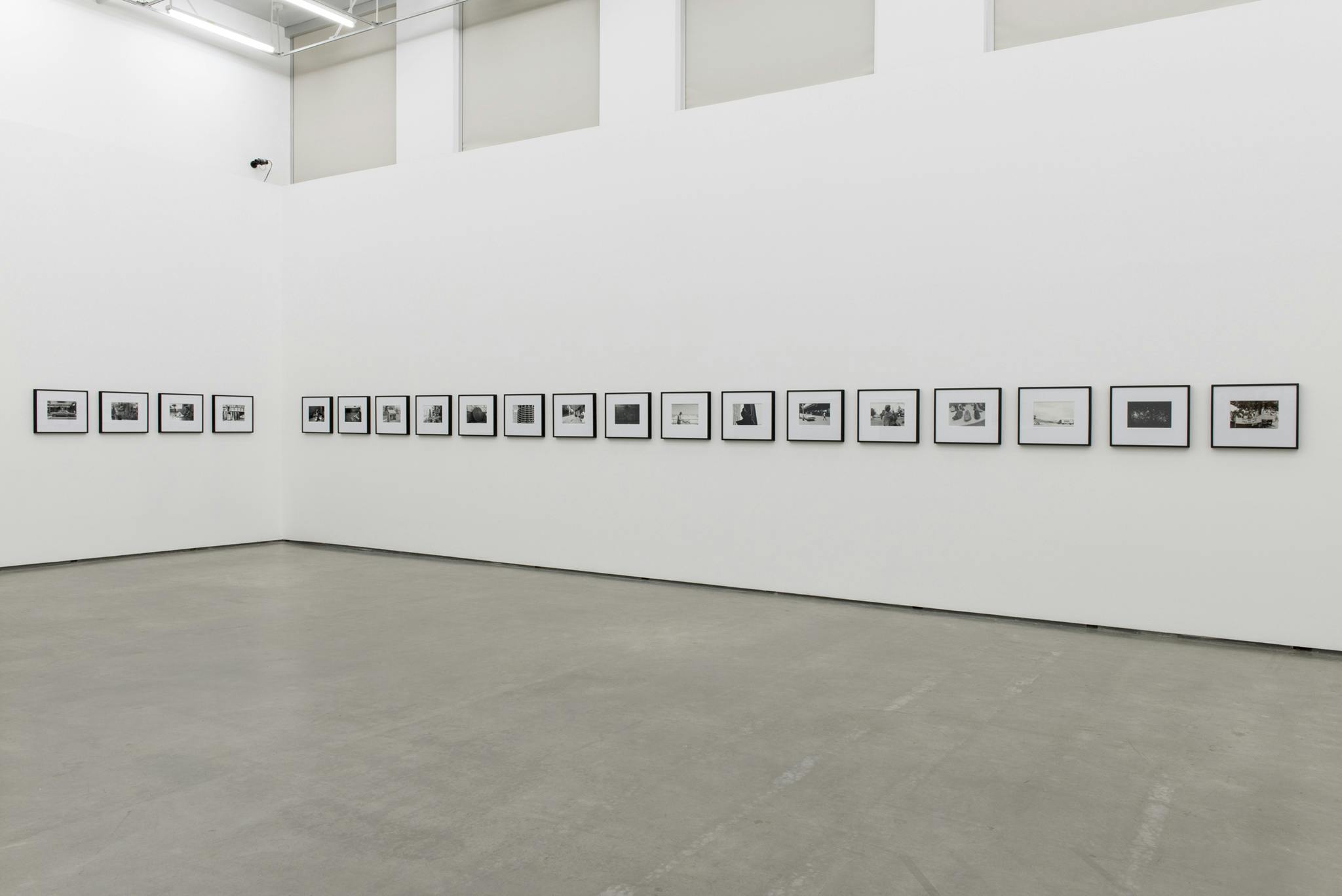 Twenty framed black and white photographs hang on two sides of a corner wall of a gallery. Four of the photographs hang on the left side of the corner wall and sixteen hang on the right side.
