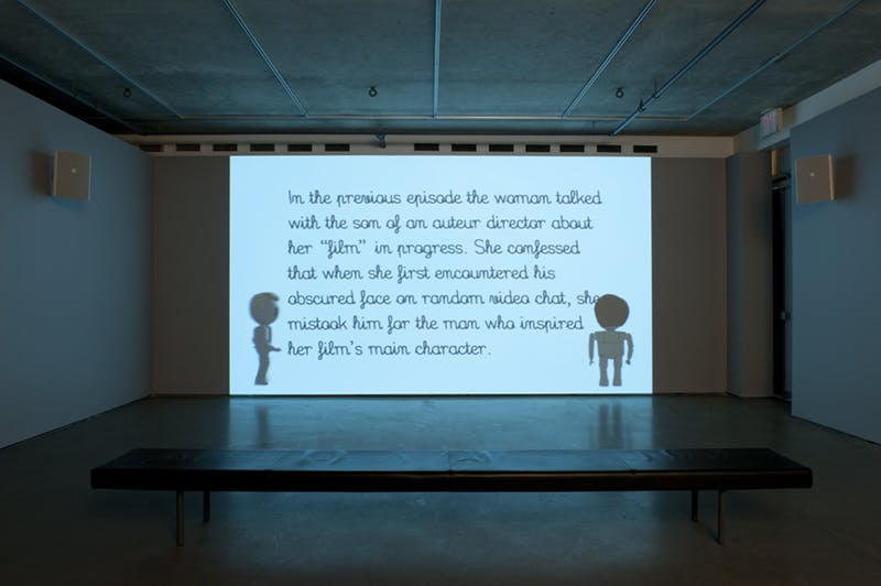 A single-channel animated video is projected on a wall in a darkened gallery space. The screen displays a synopsis of the previous episode between two naked, toy-like human figures.