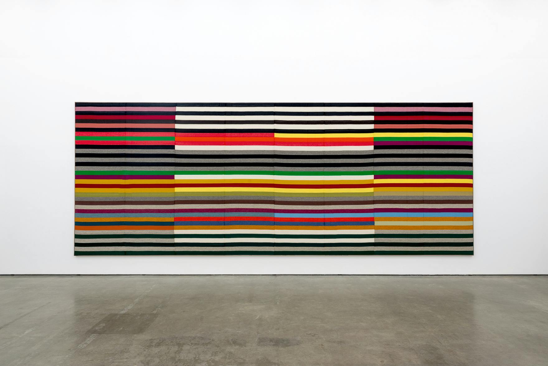 A large-scale woven piece hangs on the wall of a gallery space. The piece is rectangular and made up of various coloured and white stripes. 