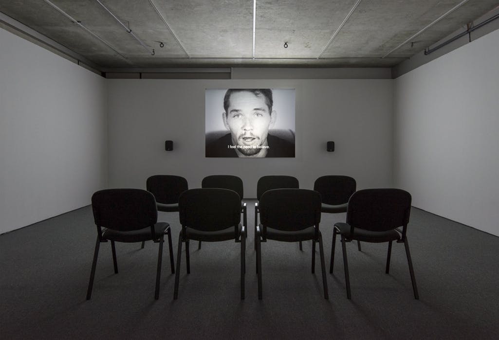 A single-channel video is projected on the wall. The video depicts a person speaking directly to the camera. The subtitle reads, “I feel the need to believe.” Black chairs are aligned on the floor.