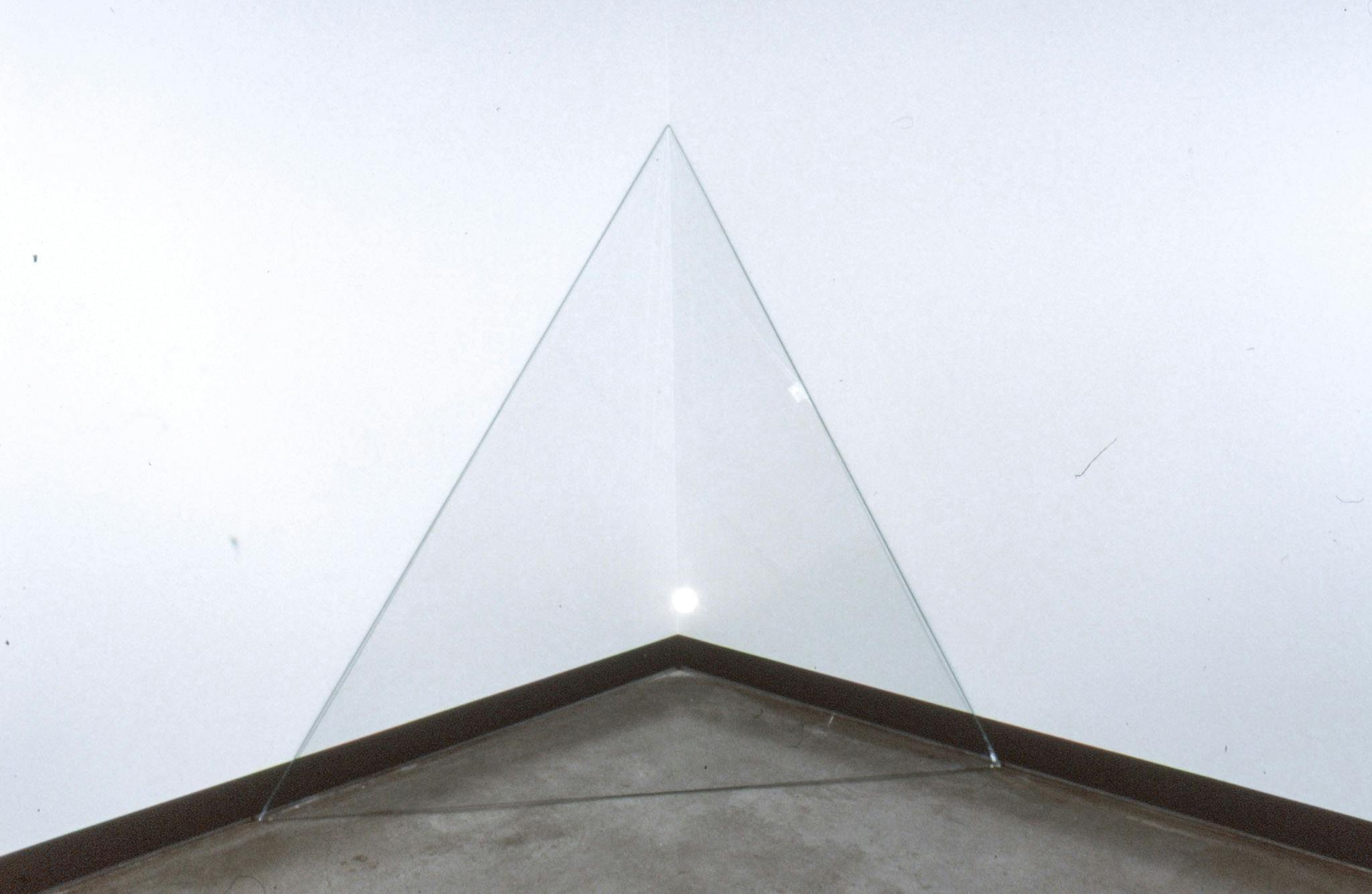 Installation image of Nestor Krüger’s artwork. A transparent board, which is cut out in a triangular shape, leans against a corner of a gallery space. The work is approximately the size of a 3-year-old child.
