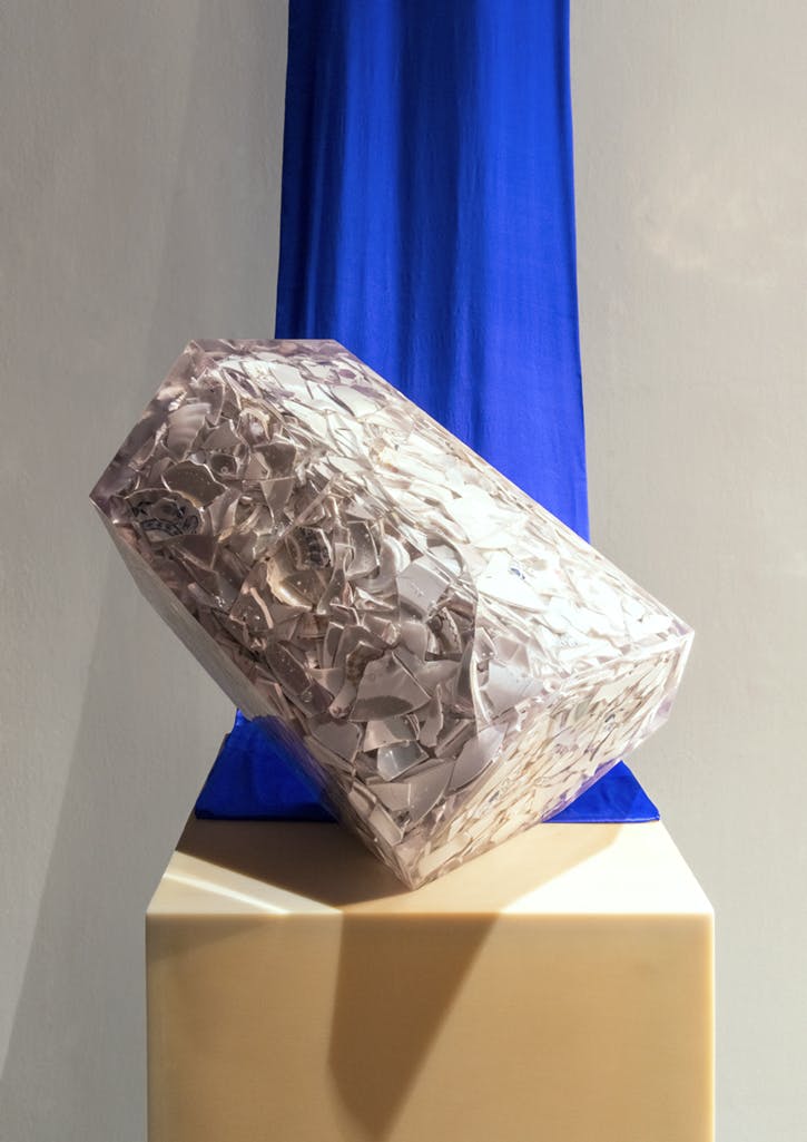A clear, block-like sculpture of encased broken ceramic pieces installed on a plinth. A royal blue cloth hangs from the ceiling and drapes behind the sculpture. 