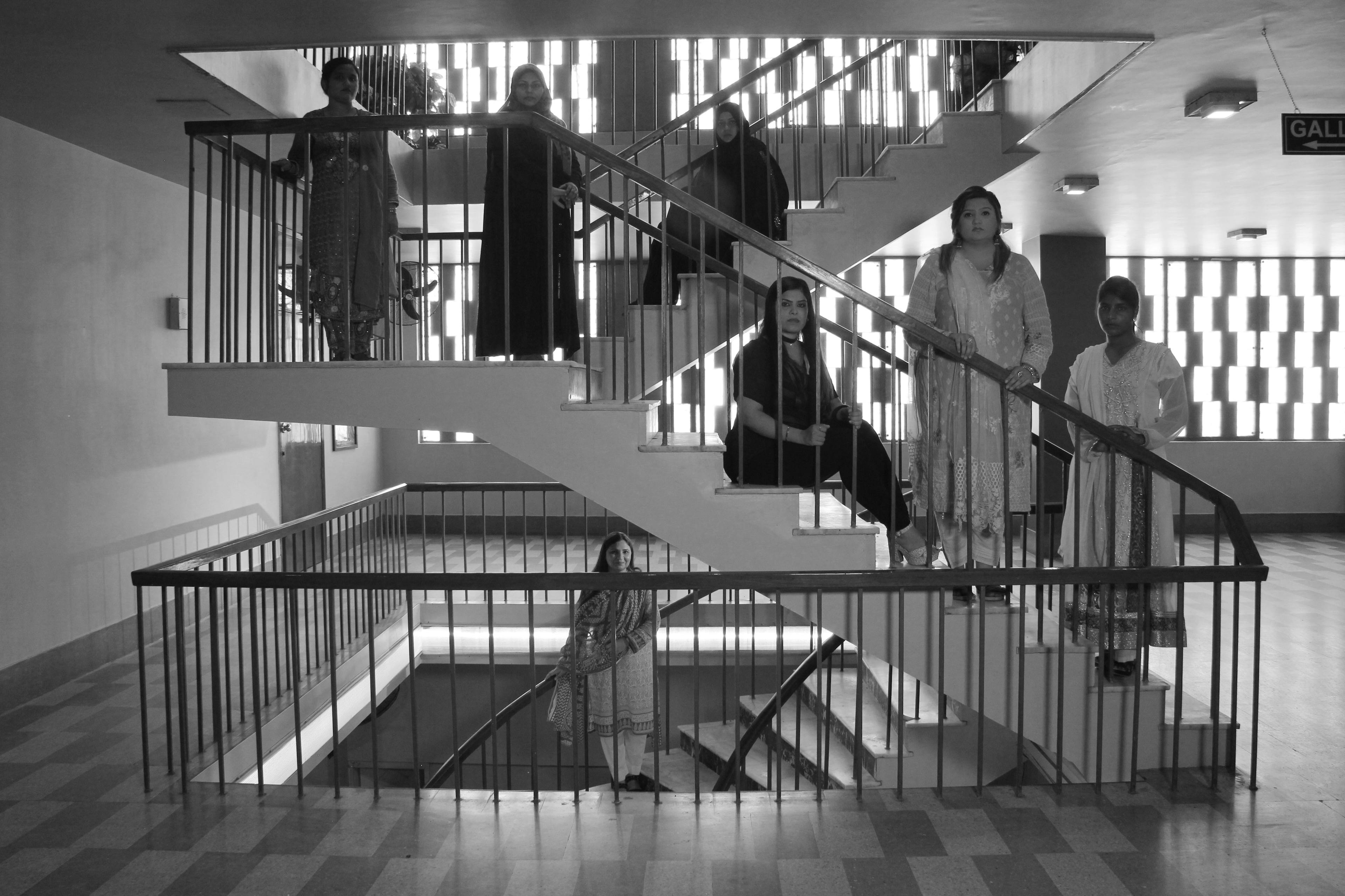 A black and white image of 7 people standing on the steps and landings of a u-shaped staircase inside of a building. Each person is looking directly at the camera. 