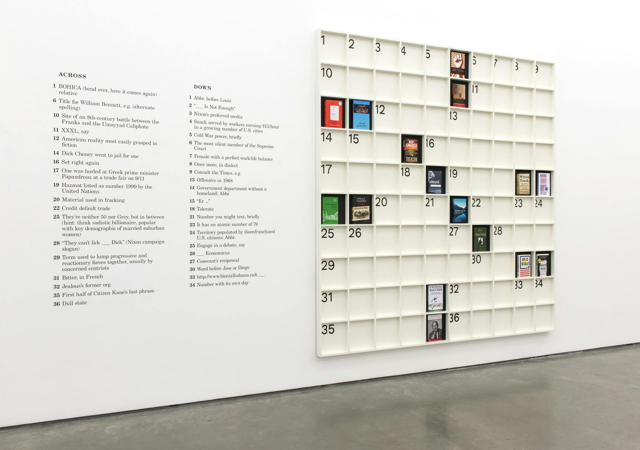 A sculpture forming a large-scale crossword puzzle hangs on the gallery wall. The dead squares of the crossword are filled with physical books affixed directly to the wooden surface. 