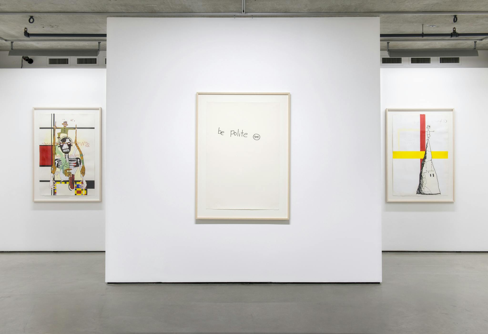 Image of a freestanding wall in front of another gallery wall. On the center wall hangs a framed work on paper that reads “BE POLITE.” Two more framed drawings hang on the back wall on either side.
