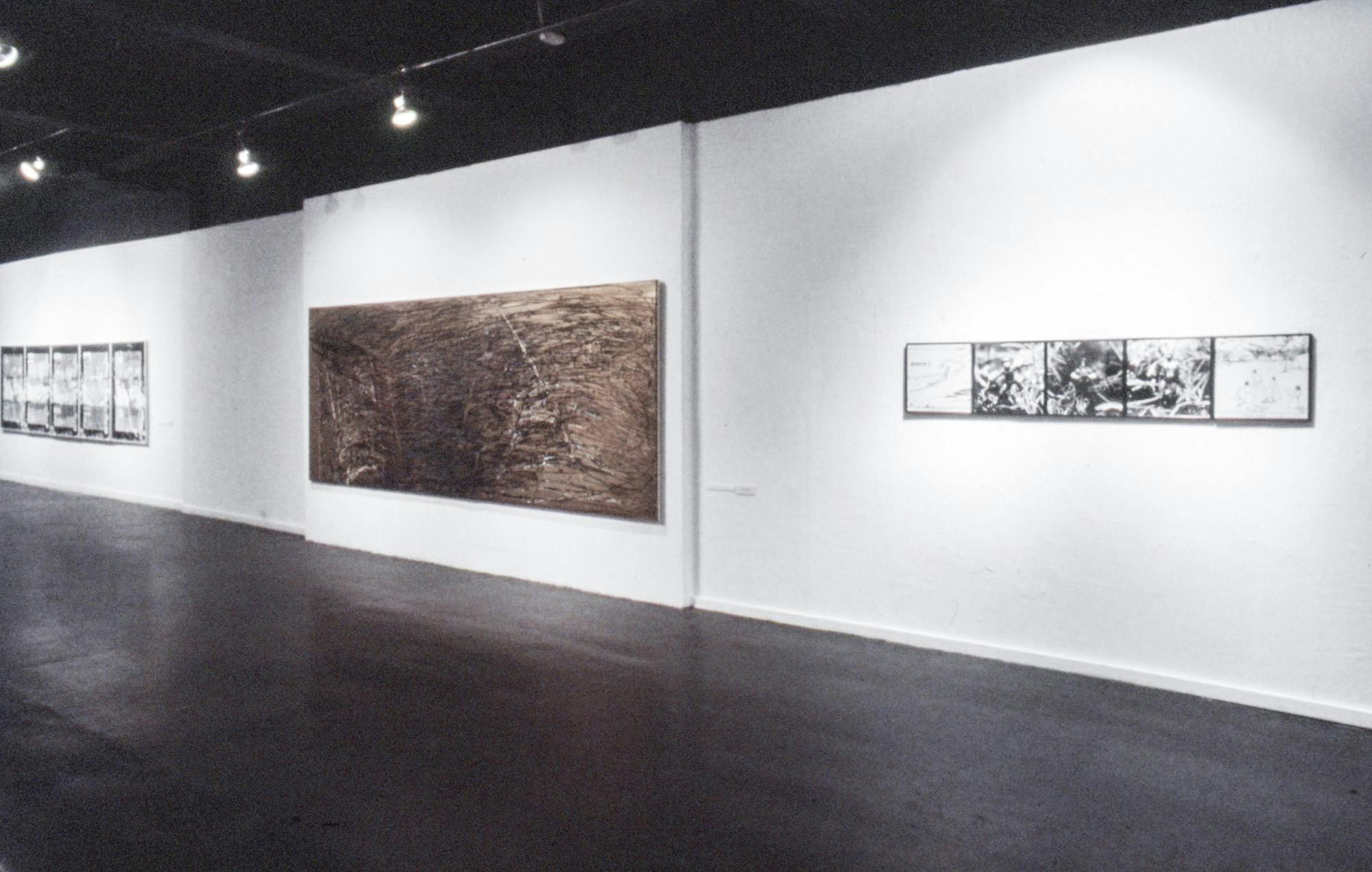 Several artworks on the wall of a gallery. At the centre there is a large abstract painting made of rough black and gold marks on canvas. On the sides, there are sets of black and white photos.