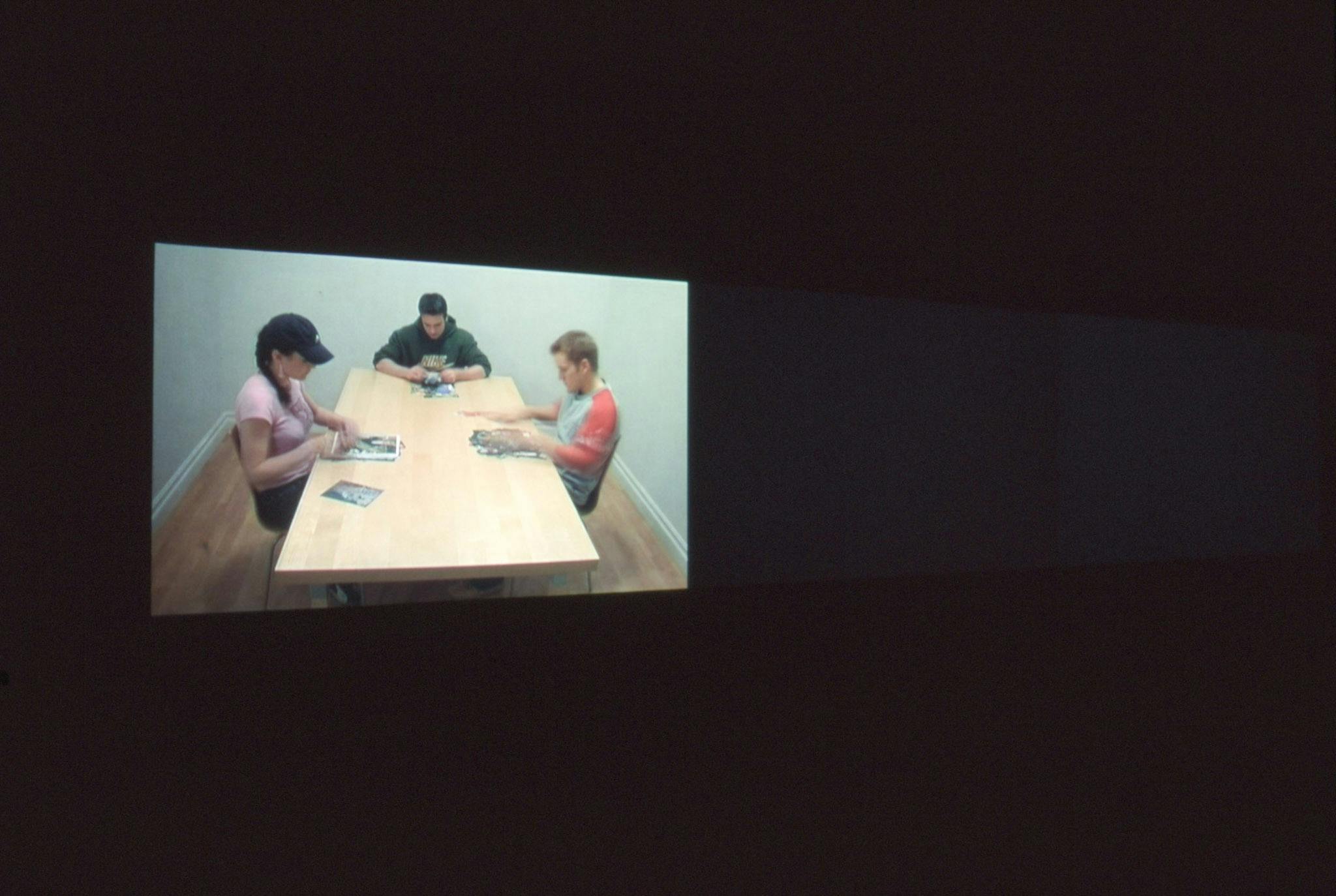 Installation image of video work by Alex Morrison. A video projected on a wall in a completely dark gallery space shows three people at a table working on stacks of magazine-like objects in front of them. 
