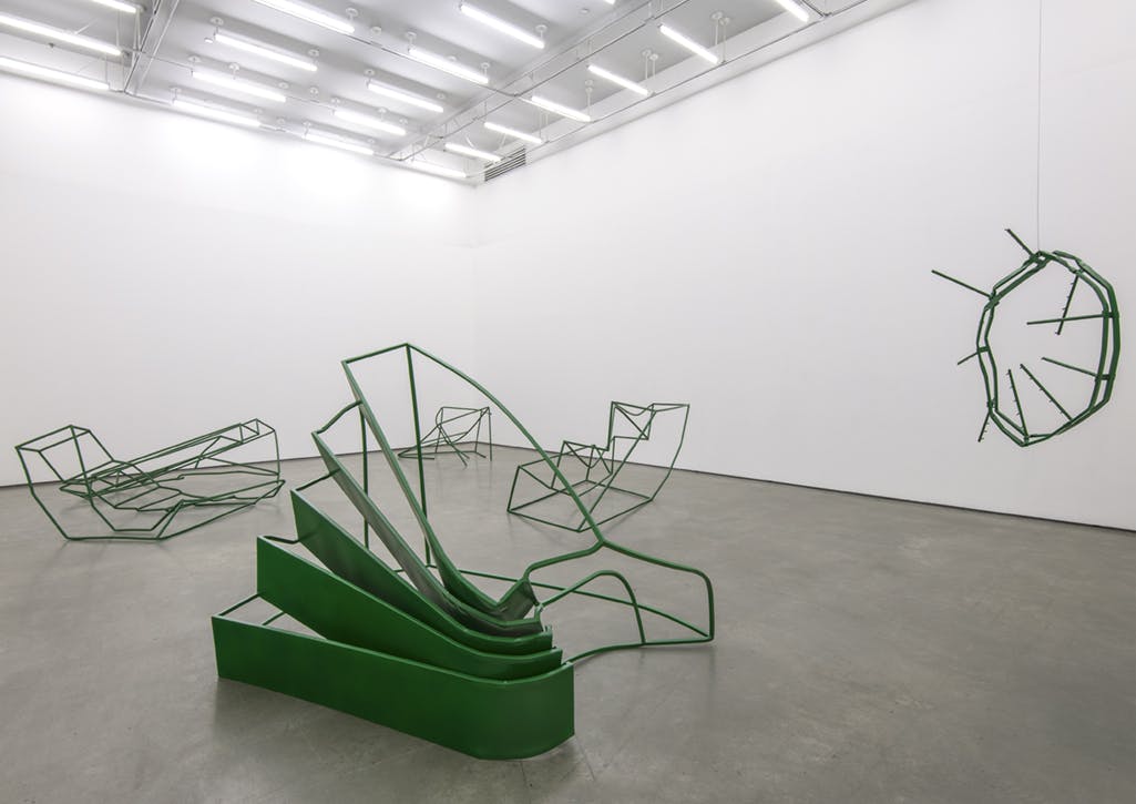 Five green, metal sculptures installed in a gallery. The sculptures resemble architectural forms that have been distorted and bent.