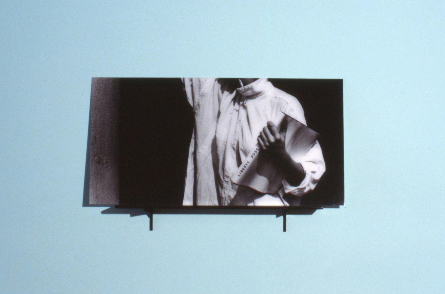 A black and white photograph mounted on a black shelf against a light blue wall. The photograph is of a person holding a book and they are only visible from the neck to the waist. 
