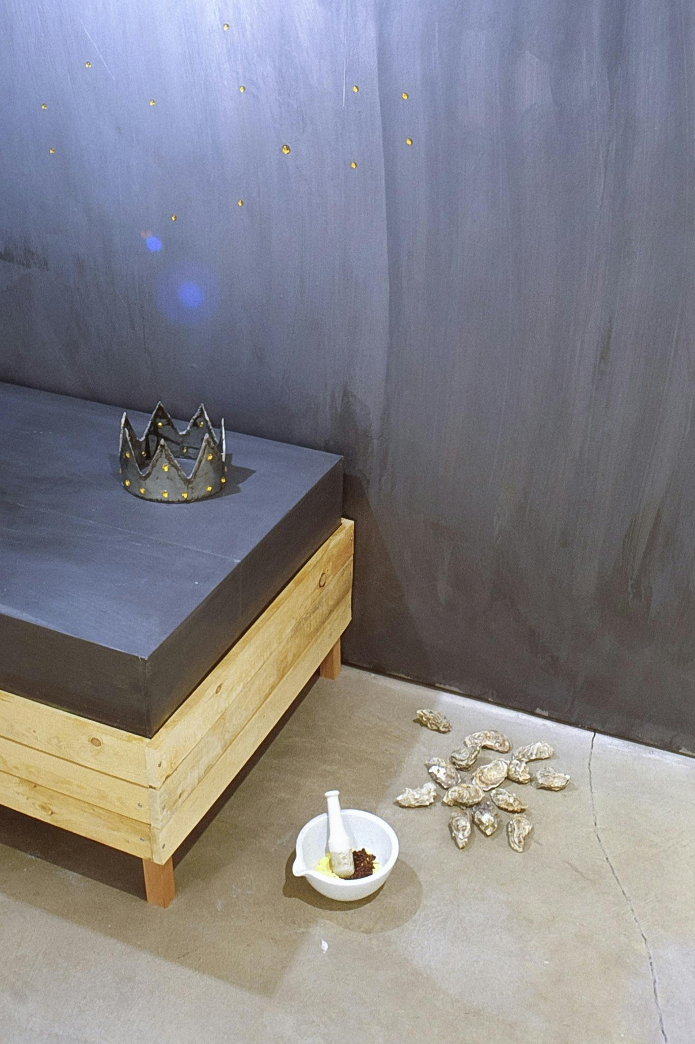 A low wooden bed frame is placed on the gallery floor, on which a silver kids crown sits. A set of white mortar and pestle and 14 oyster shells are placed on the floor next to the bed.