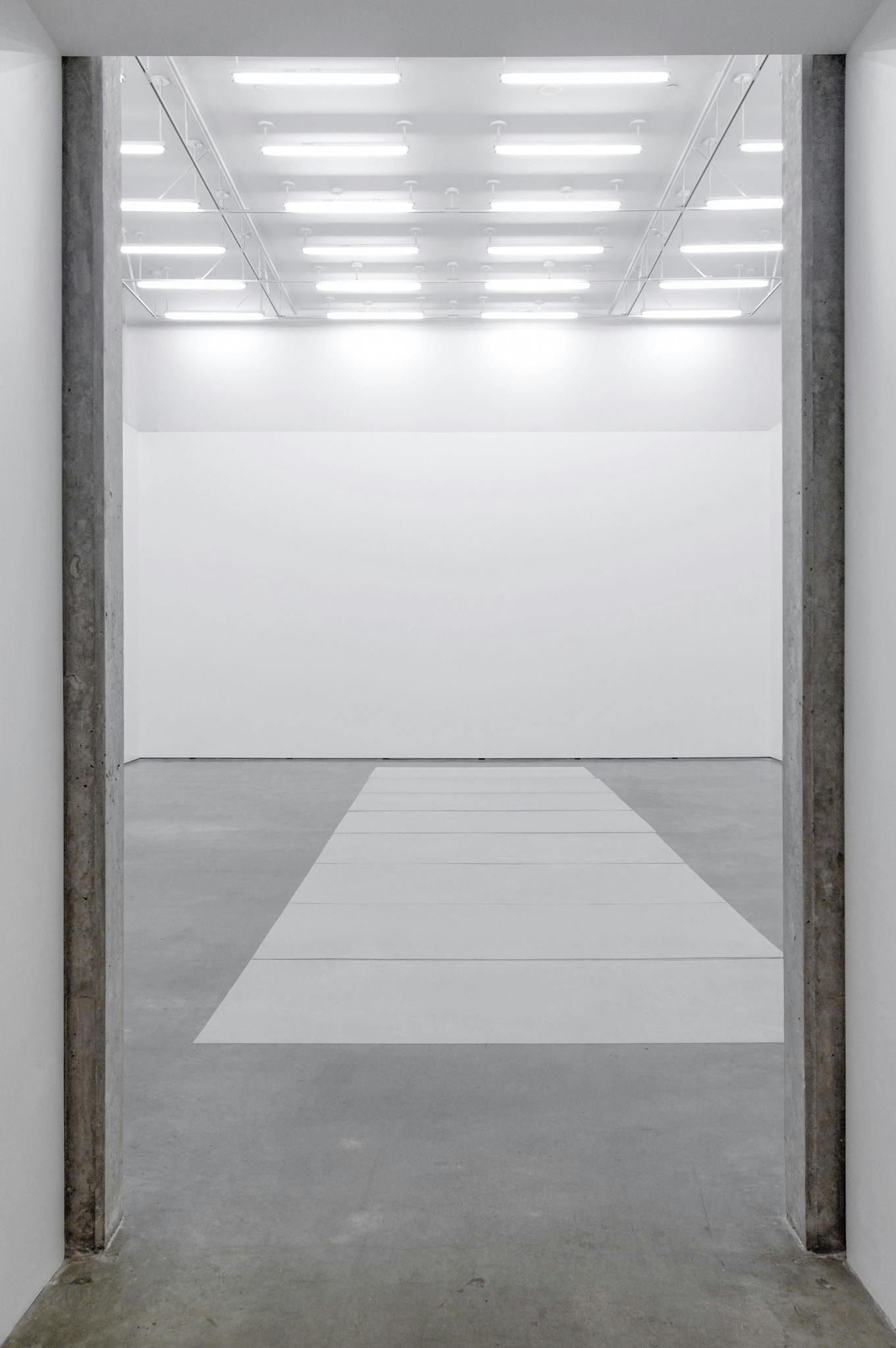 A number of rectangular-shaped mirrors are placed in a row on the gallery floor. In the same gallery space, many white fluorescent lights are installed in rows on the ceiling. 
