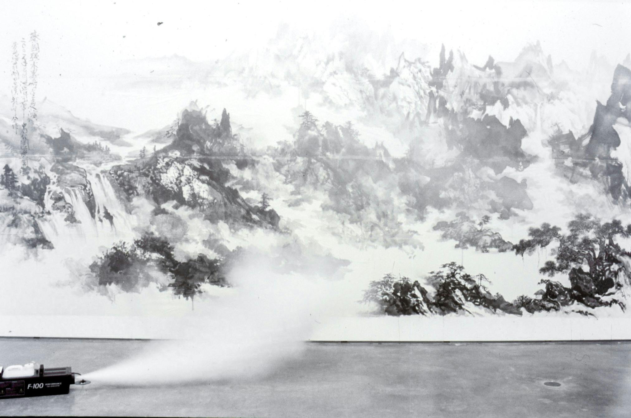 A large black ink landscape painting is mounted on a gallery wall. It shows an aerial view of mountain tops. A small machine placed on the floor emits white mist to the front of this paintings.