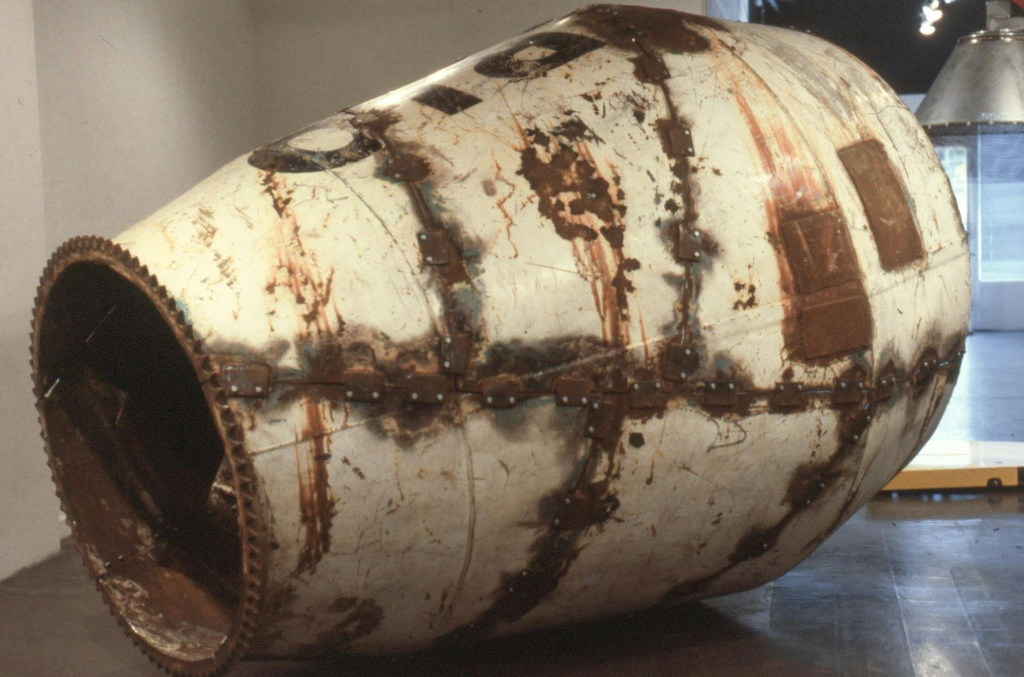 A sculpture made of a deteriorated cement mixer rests on the floor of a gallery. The exterior is white with rusty metal patches and bolts over the seams, and the interior is entirely rust coloured.