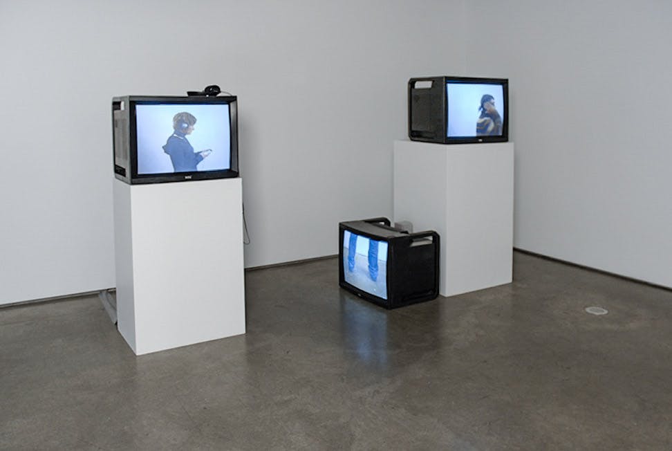 Three TV's in the corner of a galley. Two of the TV's rest on tall white plinths. The third rests on the floor, against the plinth on the right. They all play different videos of a lone person.