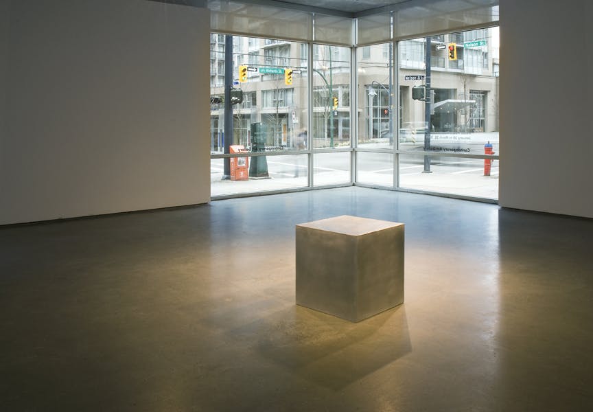 A cube sits in the middle of the gallery space. The cube is sand coloured and its size is about toddlers height. The glass window at the corner of the room shows the outside street scenery. 