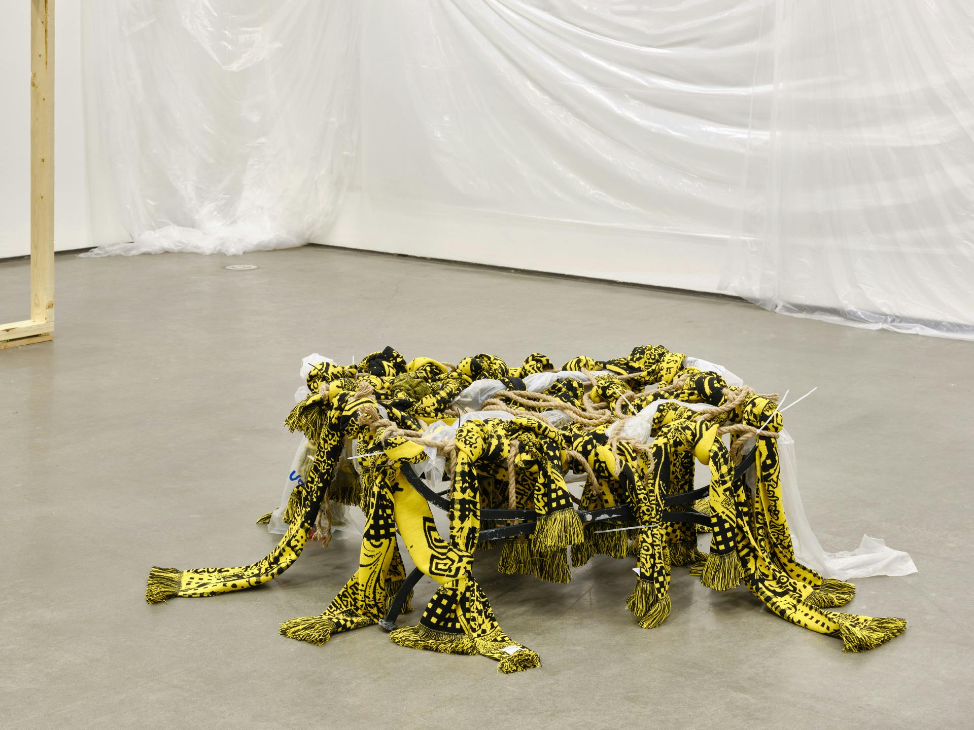  A low-lying sculpture sits on a gallery floor. The sculpture is made of yellow and black scarves, brown rope and scraps of plastic packaging tied in knots and draped over a metal seat like structure. 