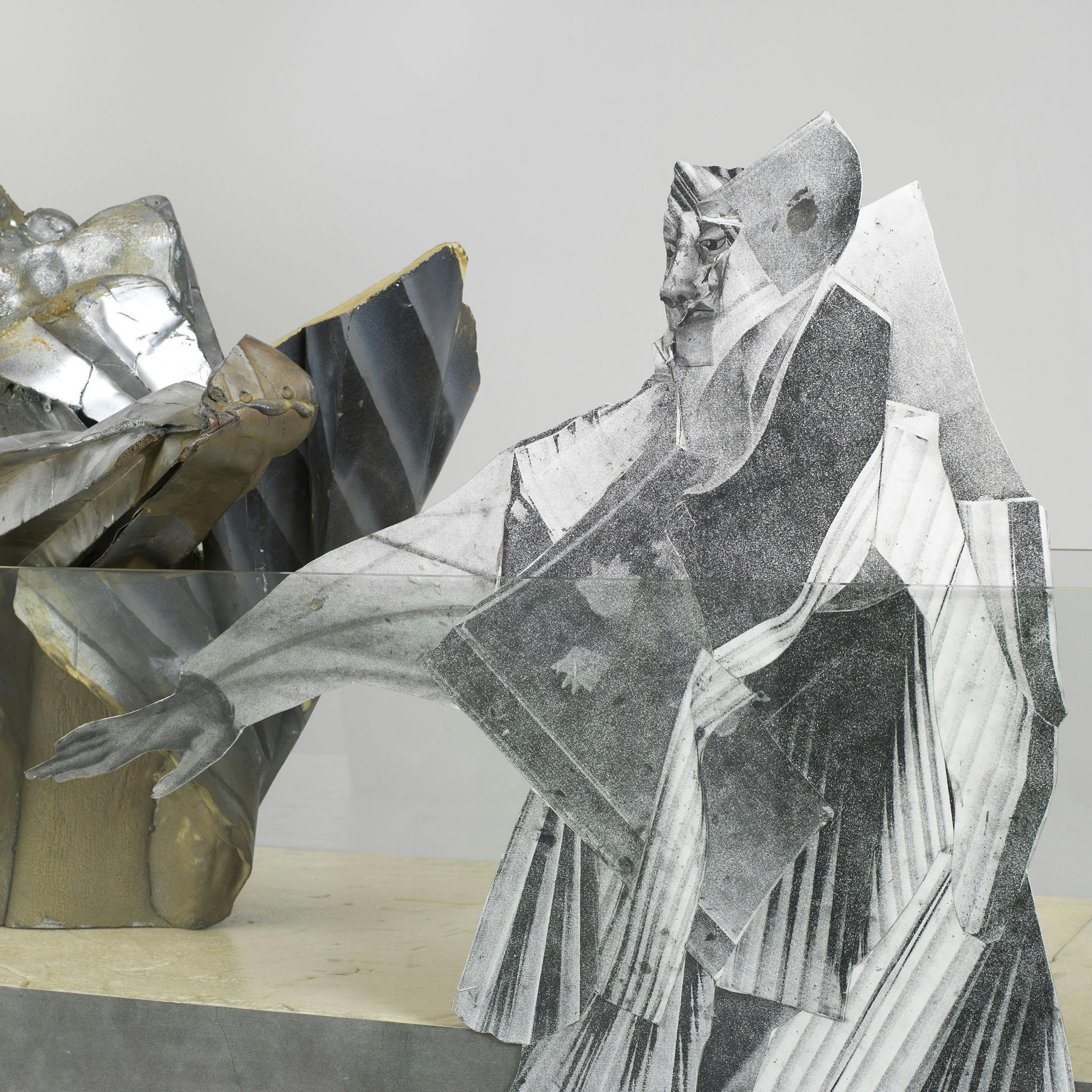 Detail image of a sculpture. A folded, collaged and photocopied drawing resembling a human figure sits on a plinth. In front of it is a sheet of glass and behind an abstract, metallic form.