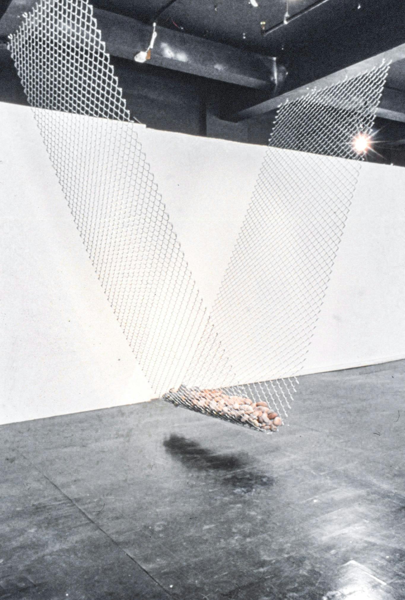 A closeup of an artwork in a gallery. The work is a large metal net suspended from the ceiling, with a row of small grey, white, and brown rocks resting in its middle.