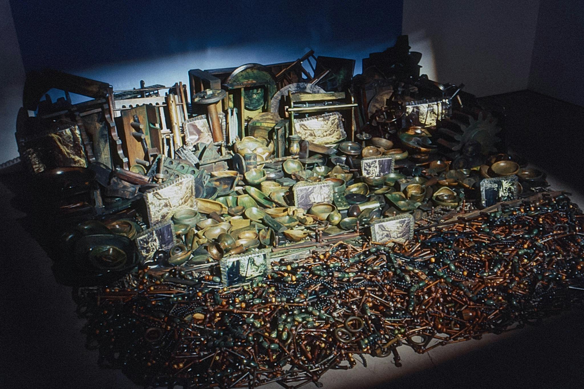 A closeup of an artwork at the base of a wall. The work is composed of hundreds of discarded objects, including framed photos, ceramic and wooden bowls, beads, industrial gears, and wall shelves.