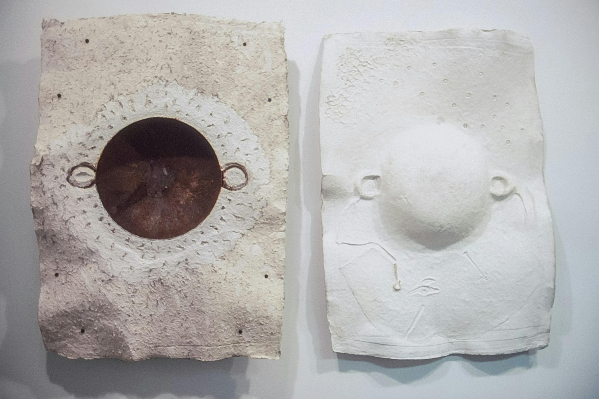 A close-up view of a pair of two artworks installed on the wall. A white pulp sheet on the right is moulded out of the round metal pan embedded in the sand-coloured rough pulp sheet. 