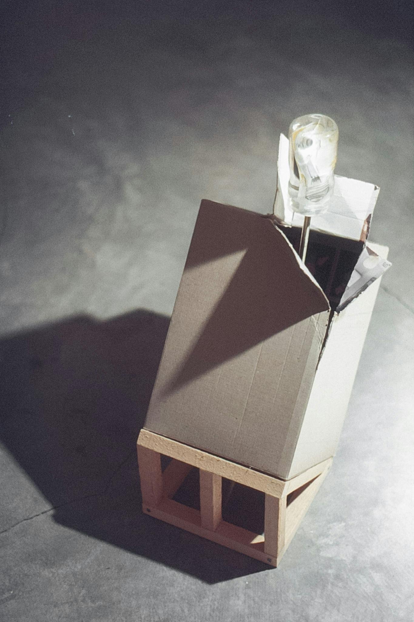 A closeup of a small sculpture on a concrete floor. The work is a cardboard box on a small wood ramp. The corner of the box is open, a screwdriver with a clear handle shaped like a lightbulb inside.