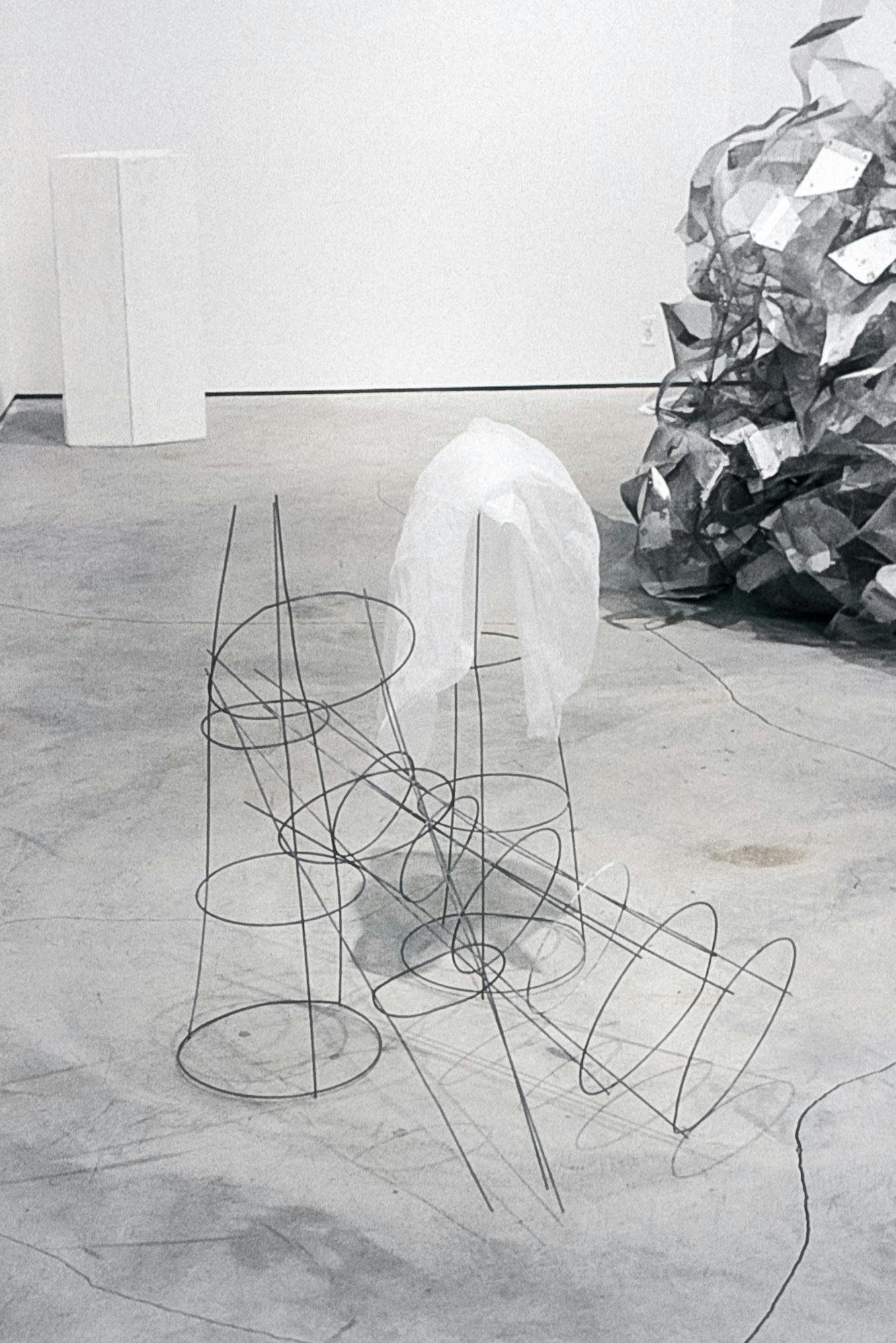 3 artworks visible in the corner of a gallery. The nearest work shows sculptures made of lines and circles of rebar wire, arranged and leaning on one another, with a piece of white fabric.
