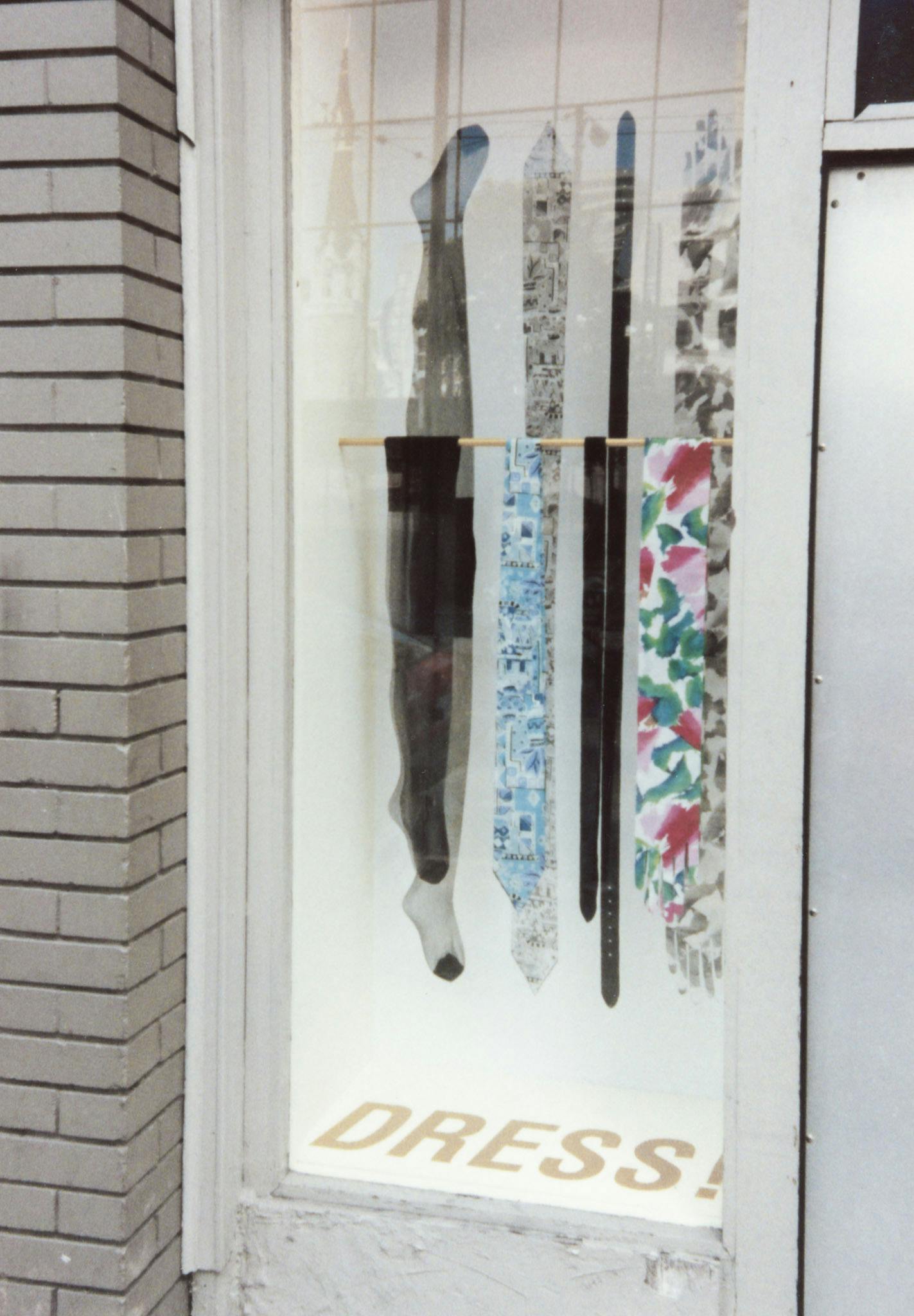 A closeup on an artwork in a window display. The work has two ties, a pair of pantyhose and a belt hanging from a rod. A photo of these items in the background. Large text on the bottom reads "Dress!"