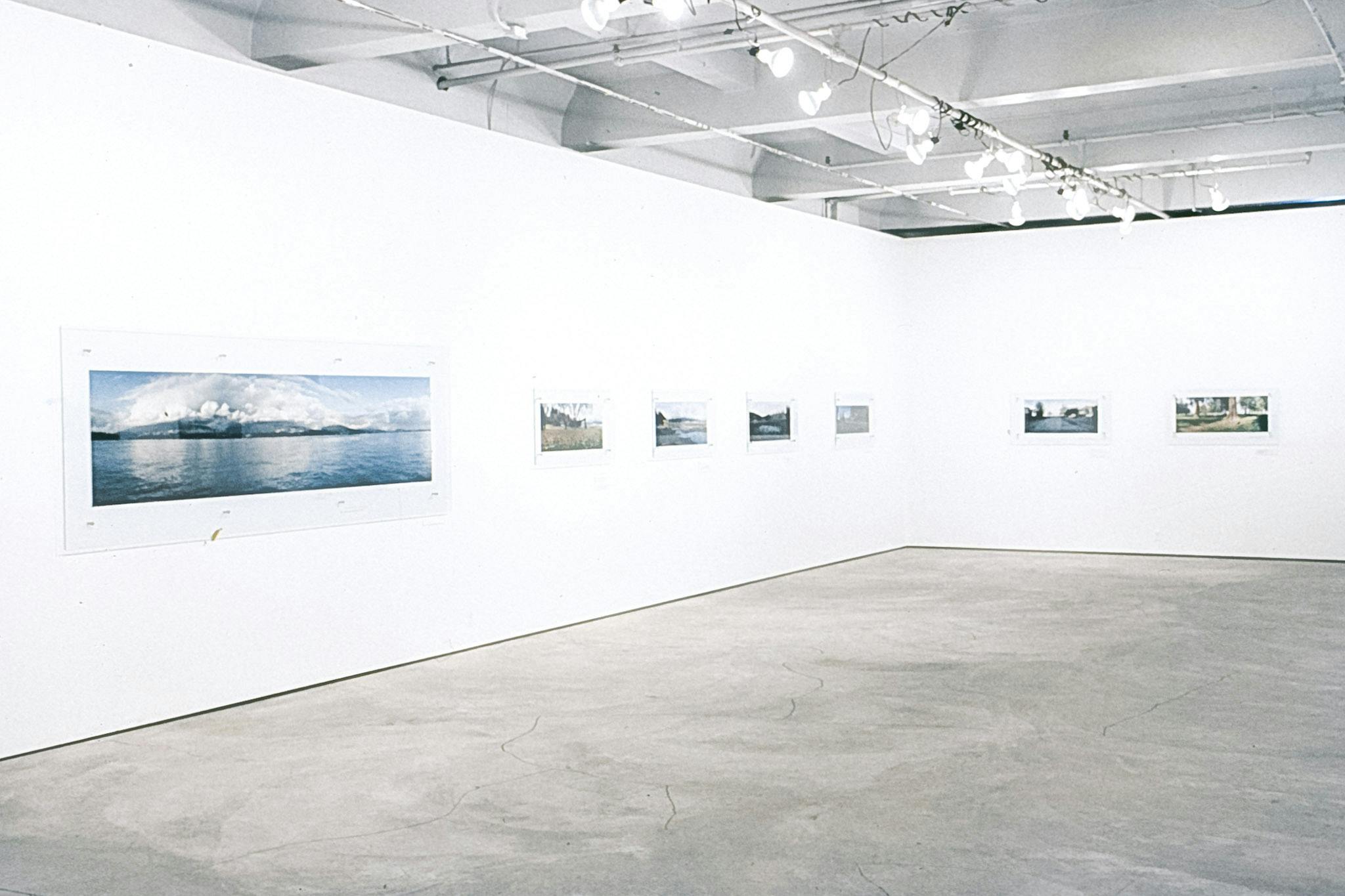Multiple photos between sheets of glass mounted on 2 white walls. The photos vary in scale and show different landscapes and places across what is colonially known as the city of Vancouver.