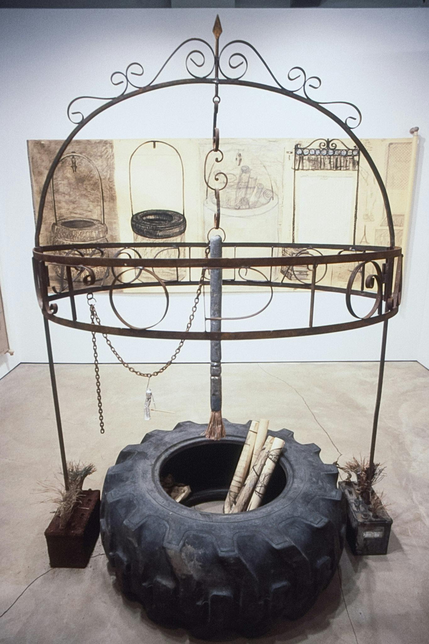 A sculpture on the floor consists of a metal gazebo, a car tire, and a large painting brush. A large black and white drawing in its back shows the design process of this architectural sculpture. 