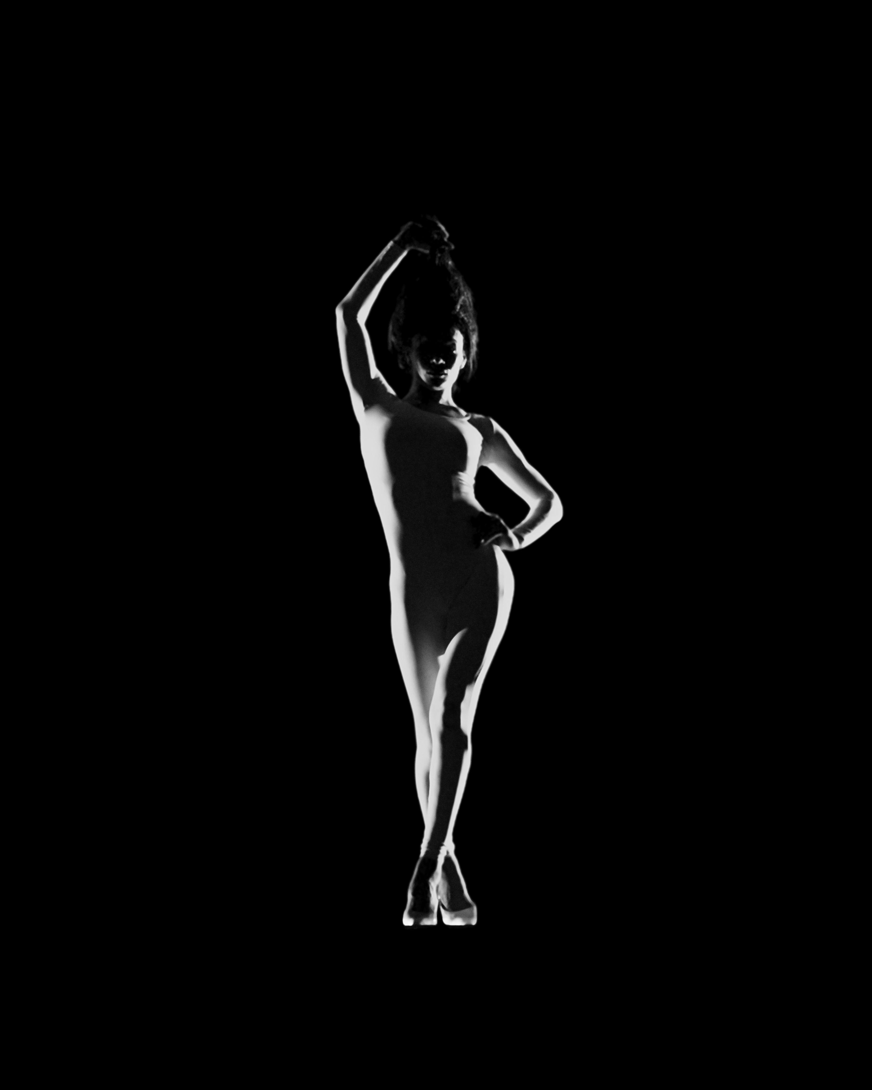 A black and white image of a figure wearing a unitard posing with one hand raising their hair above their head. the other on their hip. They are backlit and appear to be glowing on a black backdrop.  