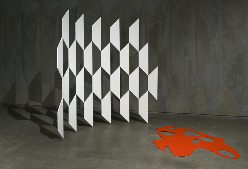 A flat, abstract-shaped sculpture in bright orange is installed in a grey-painted gallery on the floor. Beside it, a thin, white sculpture of trapezoid shapes in a fence-like pattern stands to the left.