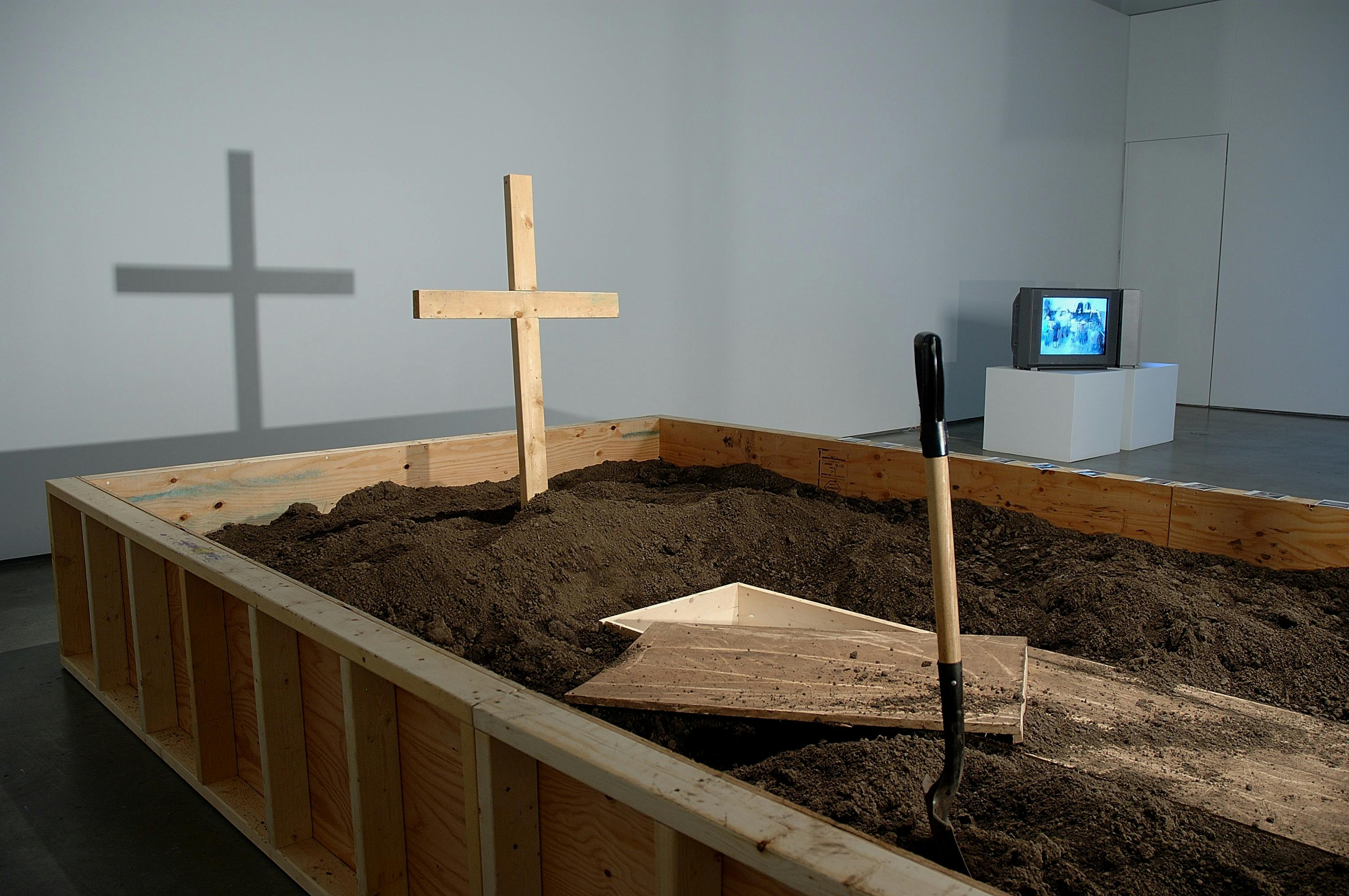 An installation image of a piece inside the gallery. A large unsealed wood box is placed directly on the floor. The box is not sealed, and a large amount of soil fills it. A cross and shovels stick out of the soil.