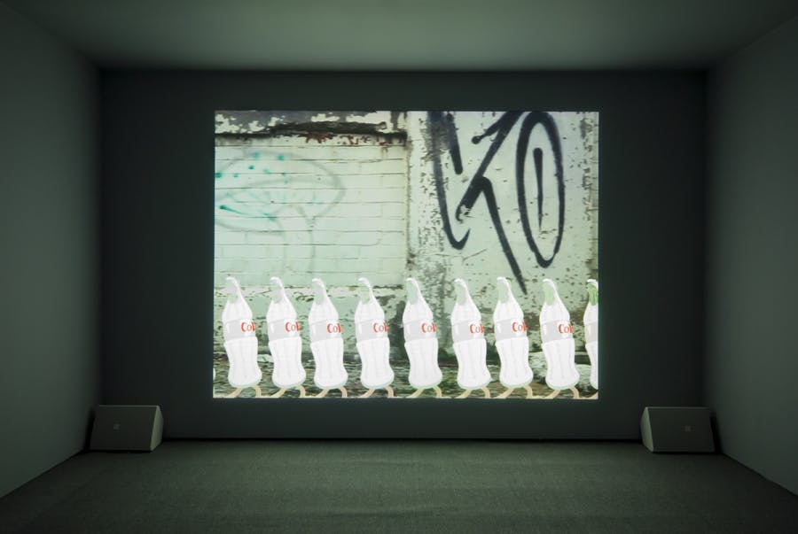 A single channel video work projected on the wall of a darkened gallery. Speakers sit on either side. The video depicts illustrated Coca Cola bottles with feet in front of a graffitied wall.