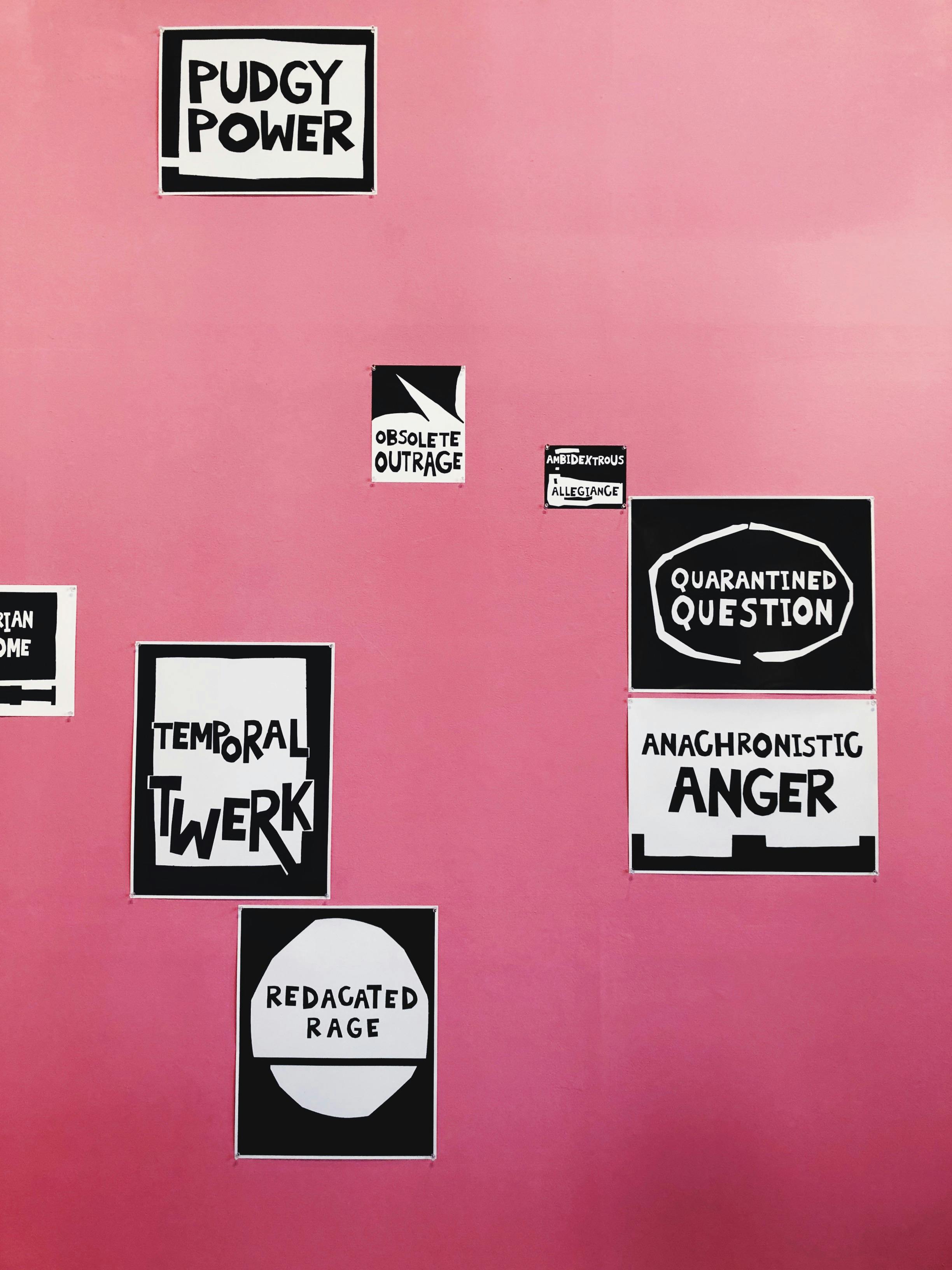 Graphic black and white text-based prints scattered across a pink backdrop. The texts are short statements such as “PUDGY POWER,” “TEMPORAL TWERK,” “ANACHRONISTIC ANGER”  and “QUARANTINED QUESTION.” 