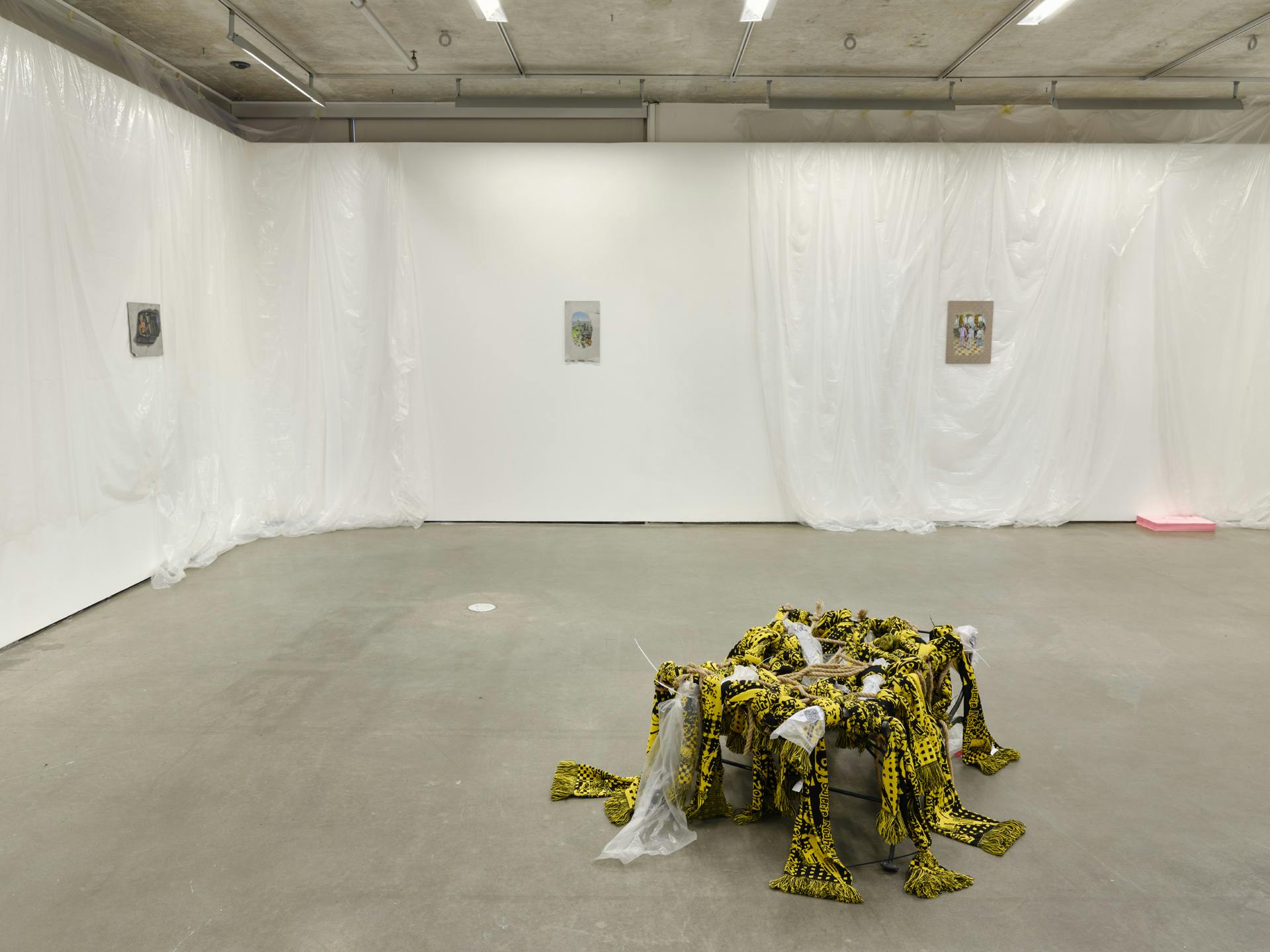  Four small paintings hang on gallery walls draped with translucent plastic sheets. A stack of pink paper and a sculpture made of yellow textiles, plastic and rope sits on the floor. 