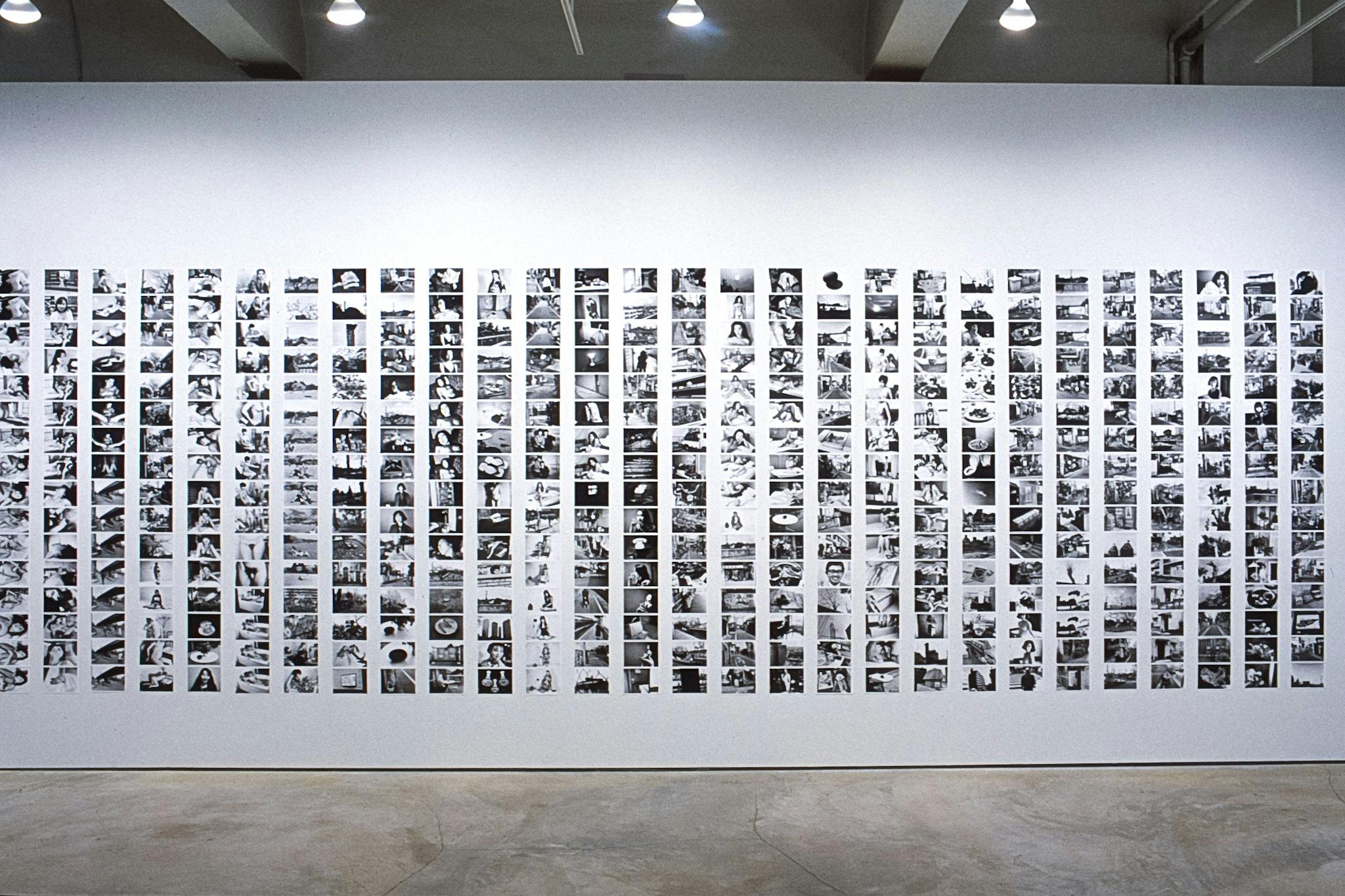 A gallery wall is filled with small black and white photographs, many of which are of a naked woman, still life, and landscape. Each vertical row has 16 photographs, and there are more than 27 rows.