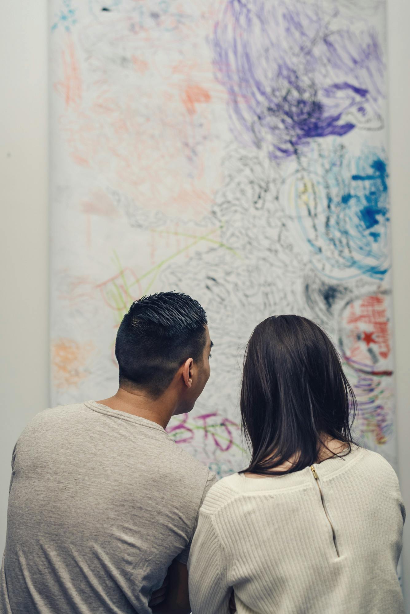 Two people, facing their back to us, look closely at a large drawing on paper, which is mounted vertically on a wall. The lines and abstract figures drawn with blue, purple, and orange markers.