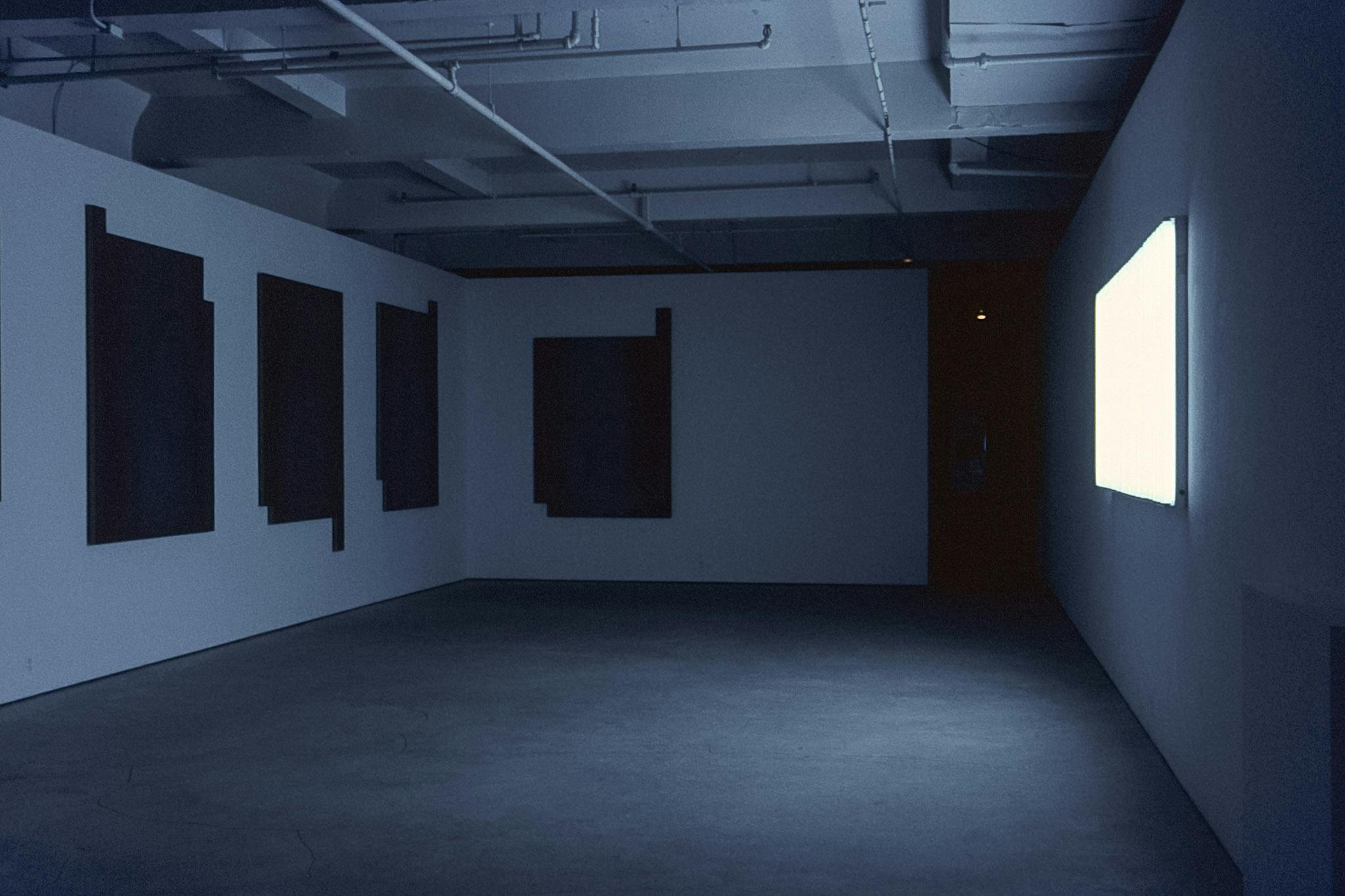 A dark gallery with several artworks  on the walls. On one wall, there is a large glowing panel. On the two other visible walls, there are several dark panels with irregular geometric corners. 