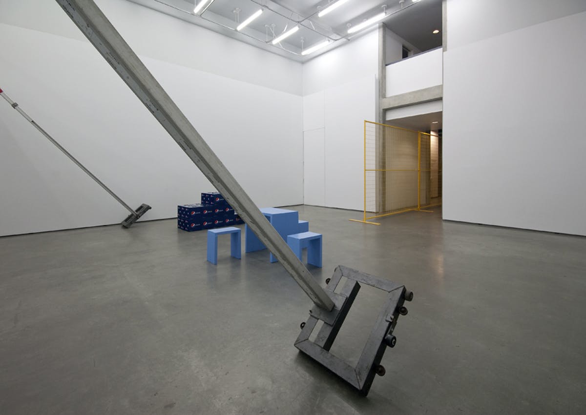 A pole vaulting stand leans against a gallery wall. On the floor, cases of Pepsi displayed as a three tier podium sit near a blue table and stools. A yellow metal fence stands in the hallway behind.