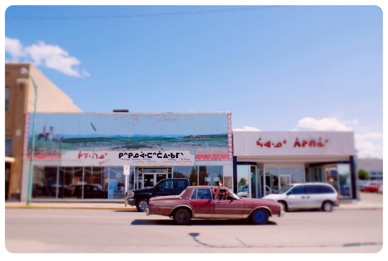 A photo with a red car driving down a street past stores and buildings, the background a clear sky. The text on the awnings has been replaced with Cree syllabics. The corners of the image are round. 
