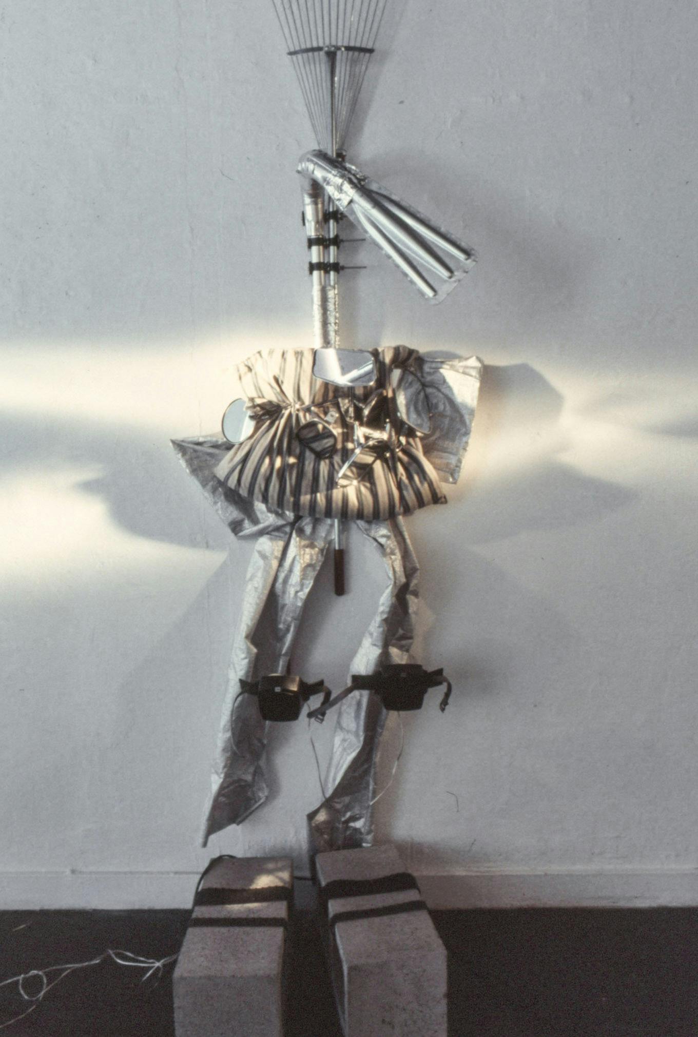 A closeup of a sculpture mounted on a wall. The sculpture is made of various readymade elements, including: cement bricks, tin foil, wires, side mirrors, metal tubes, and a rake. 