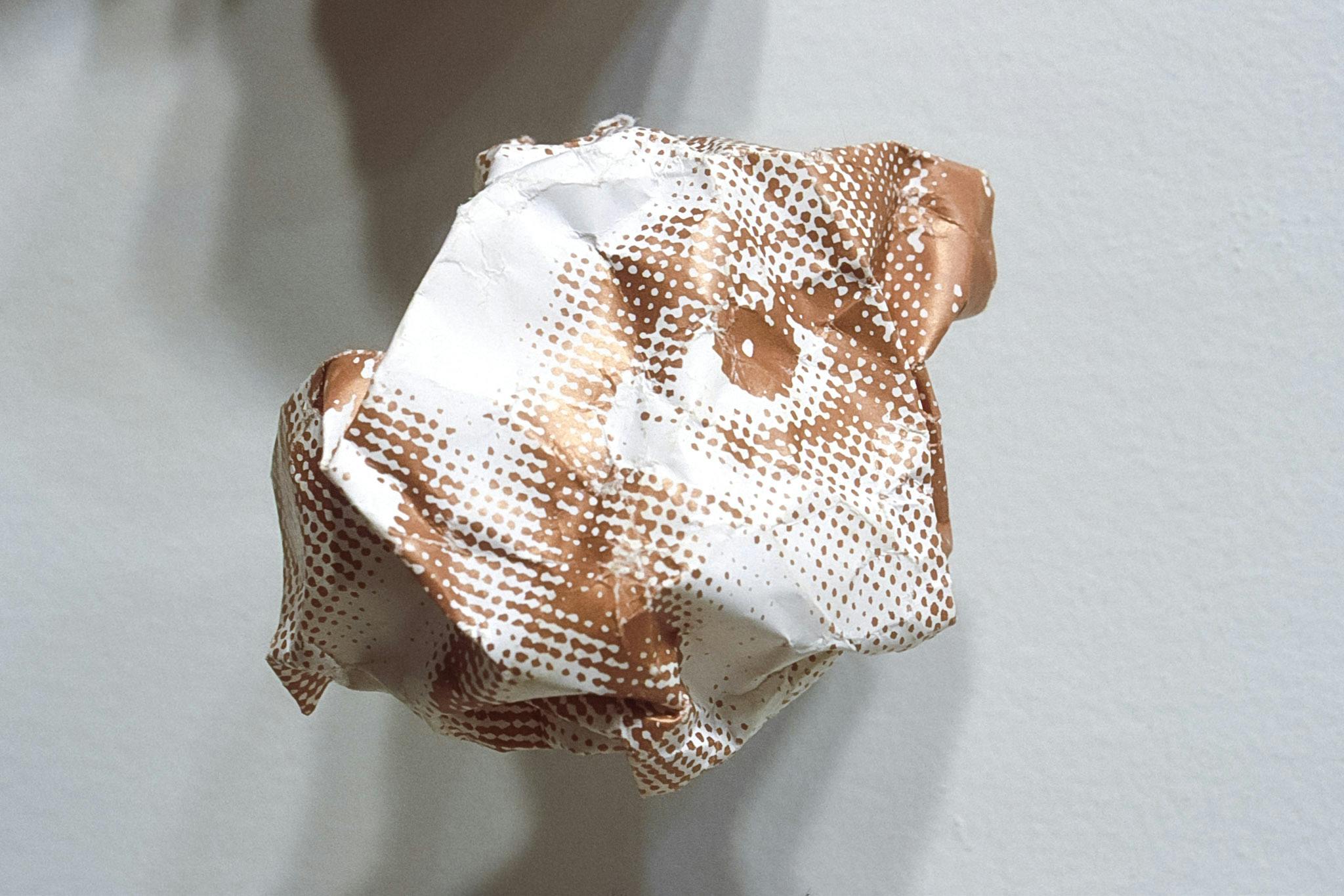 A face, printed in white and orange-brown, is roughly folded into a shape of a ball. This folded paper is installed in a gallery space. A person’s eye is the most recognizable part of this piece.