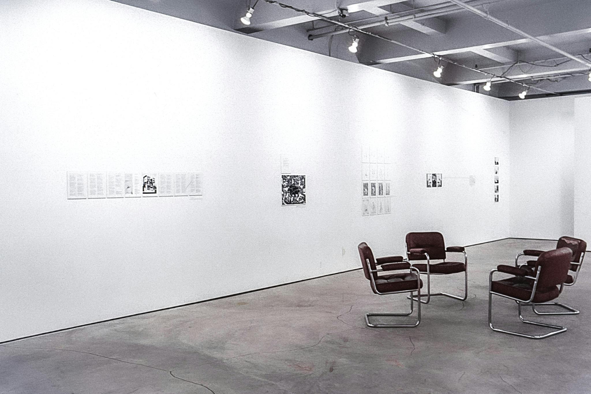 In the centre of a gallery, there are four metal and leather chairs facing each other. On the walls, there are four different artworks composed of black and white photos, and text. 