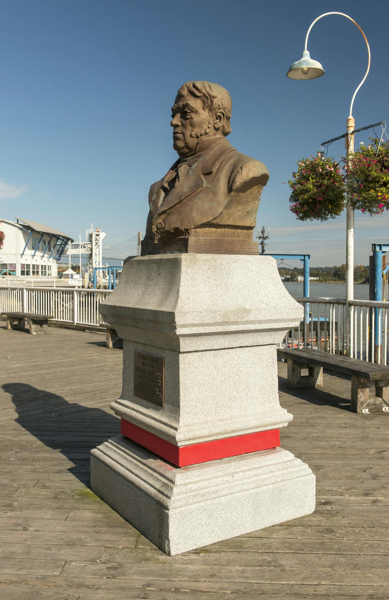 An image of a monument to Simon Fraser installed on a wooden pier by water. The monument is a bronze bust of Fraser atop a granite plinth. 