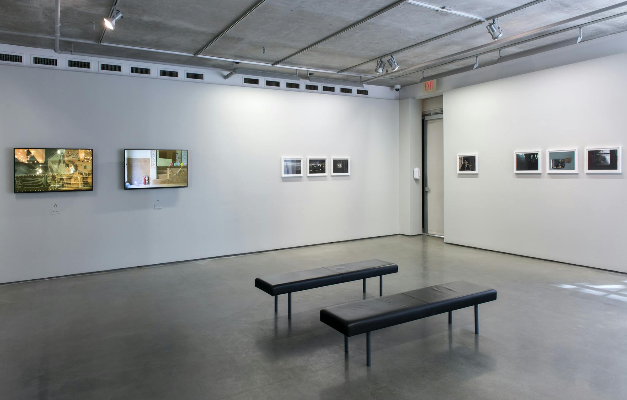 Seven framed photographs and two monitors hang on two gallery walls. The left wall has two monitors and three photographs, the right wall has four photographs. Two benches sit in front.