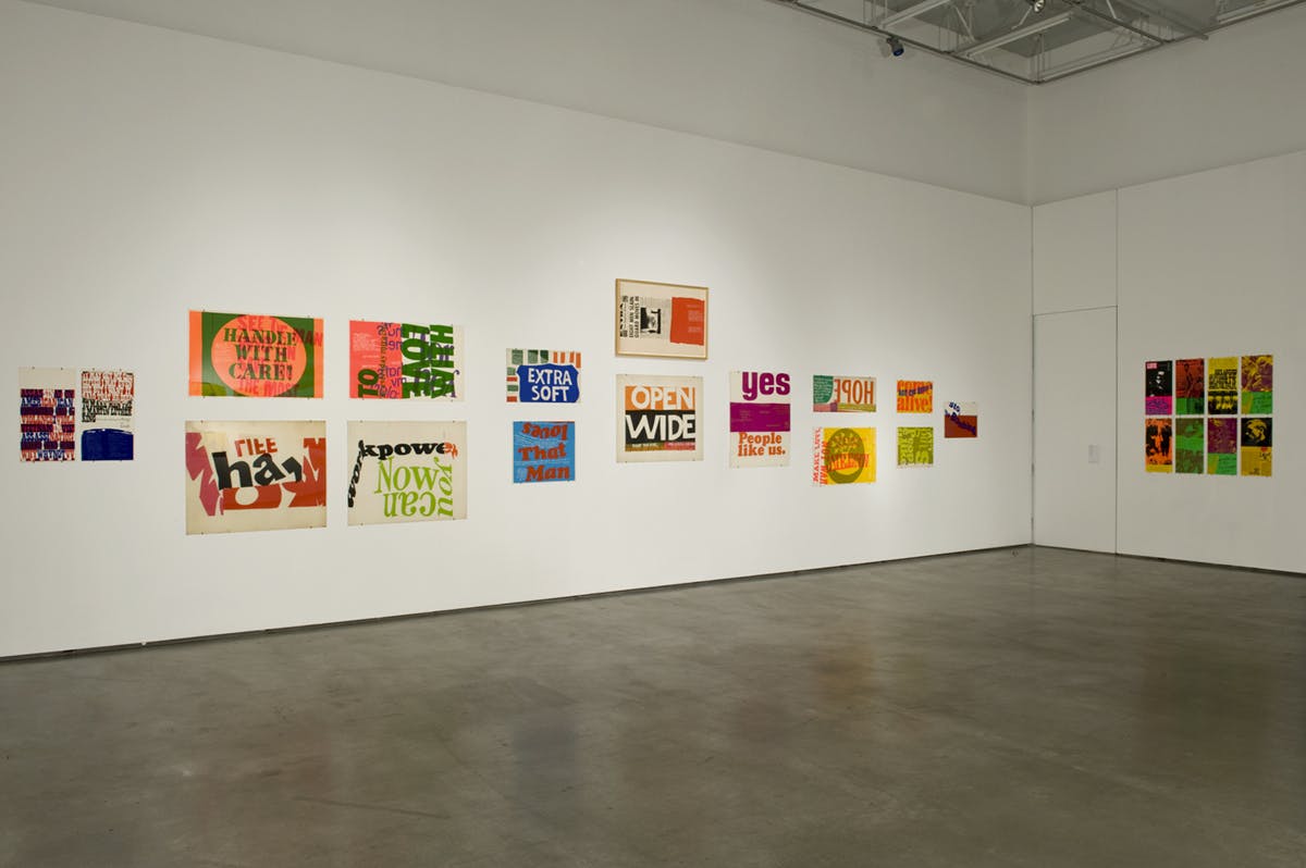 Installation view of Corita Kent’s colourful sign artworks mounted on white gallery walls. Some signs have their words mirrored, disproportionated, or overlapped with each other.