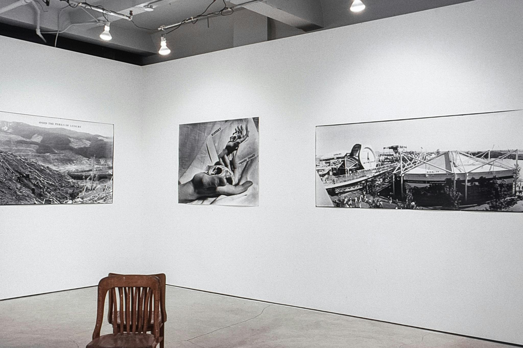 3 black and white photos on the walls of a gallery. The photos show a landscape, a collage of a hand holding a small object, and an arena. In the foreground there are 2 wooden chairs, back to back.